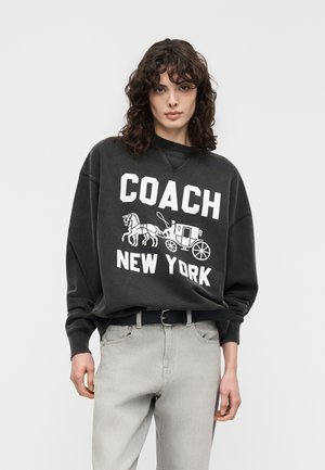 Kvinde med krøllet hår iført en sort oversized sweatshirt med hvid tekst "COACH New York" og grafik af en hestevogn, lysegrå jeans.