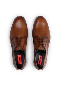 Chaussures en cuir marron avec une texture lisse, bout rond et semelles noires. Présentent un laçage et un logo rouge et noir sur la semelle intérieure.