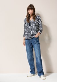 Blusa de patrón azul marino y blanca con líneas geométricas, presenta un escote en V y mangas largas. Combinada con jeans de mezclilla de pierna ancha en azul claro y zapatillas blancas.