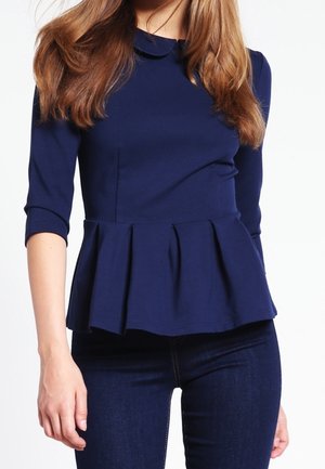 Vrouw draagt een marineblauwe peplum top met driekwart mouwen en donkerblauwe jeans, staand tegen een effen witte achtergrond.