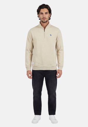 Man wearing beige quarter-zip sweatshirt, black jeans, and white sneakers, standing against a plain light background.