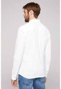Camicia bianca a maniche lunghe con colletto, caratterizzata da una texture liscia e bottoni sui polsini, abbinata a jeans azzurri chiari.