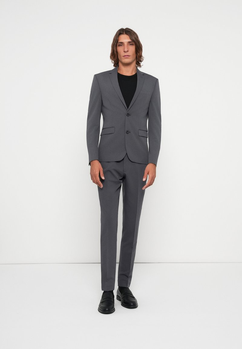 Gray suit with a fitted blazer and flat-front trousers. Features notch lapels, two buttons, and pockets. Worn over a black shirt and black shoes.