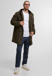 Olivgrüne Parkajacke mit fleecegefütterter Kapuze, Fronttaschen und Knopfverschluss, getragen über einem weißen T-Shirt und blauen Hosen.