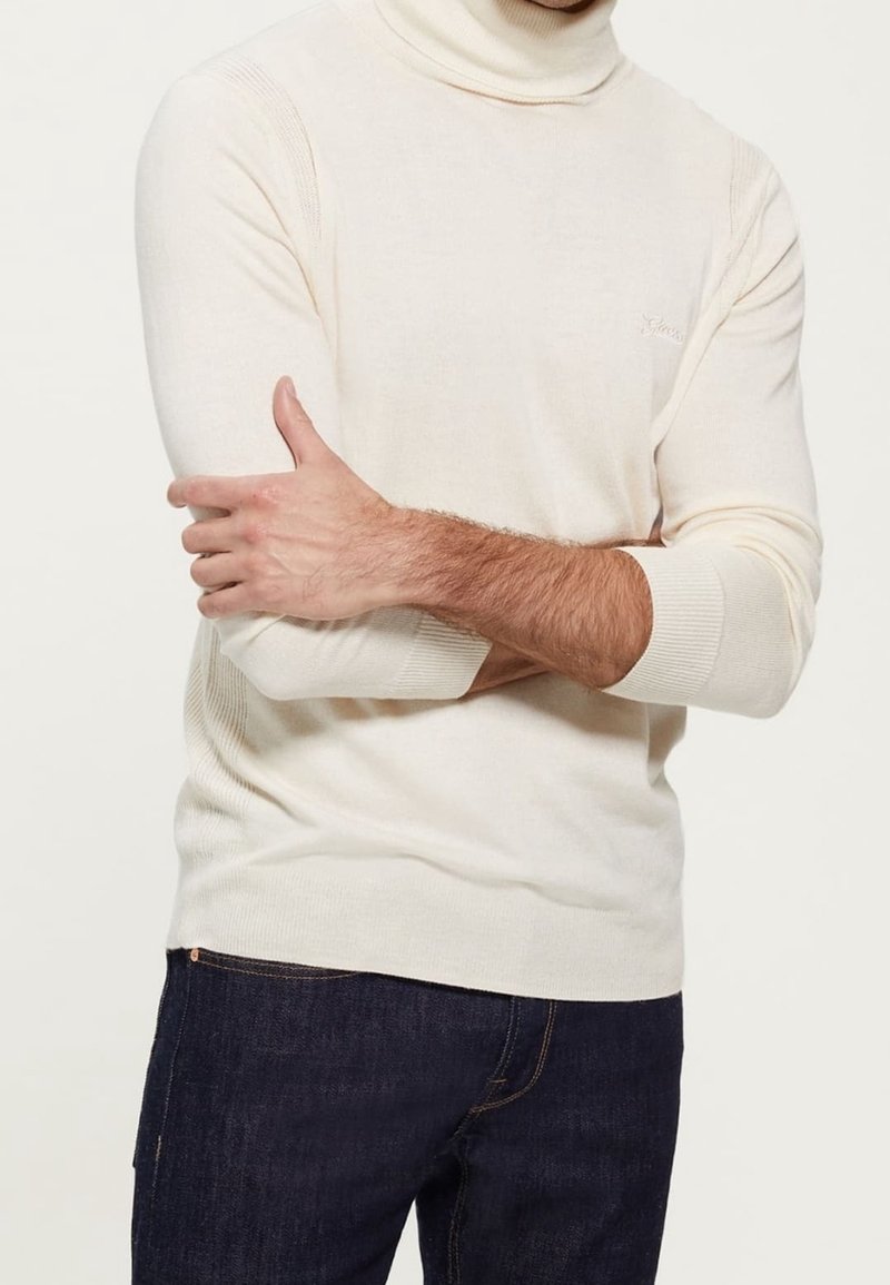 Cream turtleneck sweater in a ribbed texture with fitted sleeves, paired with dark blue jeans. Subtle branding on the chest.