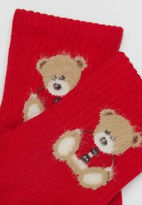 Červené pletené ponožky s motivem medvěda, medvěd má texturovaný, načechraný povrch, zatímco základ ponožky je hladce pletený.
