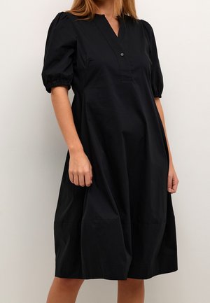 Vrouw draagt een loszittende zwarte jurk tot op de knie met pofmouwen en een V-hals met knopen op de borst.