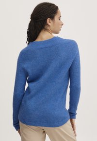 Zils, mīksts adījuma džemperis ar garām piedurknēm un noapaļotu apkakli. Tam ir teksturēta virsma un brīvā piegriezuma siluets. Nedaudz ribots apakšmalā.