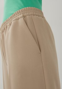 Pantalons fuselés beiges avec une taille élastique, dotés de poches latérales et d'un tissu lisse et léger, présentés avec un haut vert.