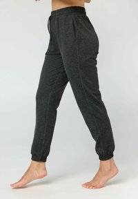 Barfußperson, die dunkelgraue Jogginghosen mit elastischen Bündchen und Bund trägt, aufgenommen im Gehen vor einem einfachen hellen Hintergrund.