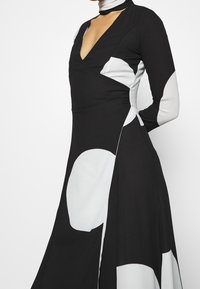 Schwarz-weißes Kleid mit langen Ärmeln, das einen tiefen V-Ausschnitt, ein asymmetrisches Design und große weiße geometrische Muster auf schwarzem Hintergrund aufweist.