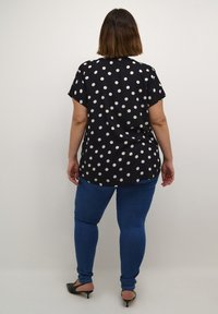 Maglietta nera con pois bianchi, maniche corte e vestibilità ampia, abbinata a jeans blu e scarpe con tacco nere. Vista dal retro.