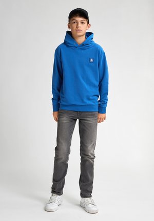 Blauwe hoodie met een logo aan de voorkant, gecombineerd met grijze spijkerbroeken en witte sneakers. De hoodie heeft een grote kangoeroezak en een verstelbare capuchon.