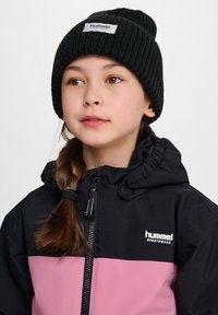 Zwarte en roze sportieve jas met een capuchon, uitgerust met een rits en logo. Draag met een zwarte geribbelde beanie. Haar in een vlecht gestyled.