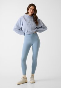 Hoodie court bleu clair avec logo brodé, assorti à des leggings taille haute et des baskets blanches avec des accents beiges.