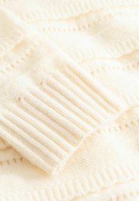 Hellcremefarbener, gestrickter Pulloverstoff mit gerippten Bündchen, der ein strukturiertes Muster und eine feine Webart aufweist. Weiches und leichtes Material.