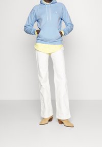 Felpa blu con lacci bianchi, indossata sopra una maglietta gialla, abbinata a pantaloni bianchi a gamba larga e stivali beige con tacco.