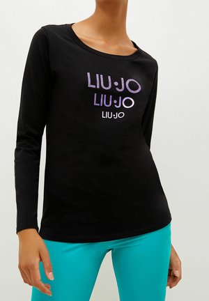 Persoon die een zwarte longsleeve draagt met het "LIU·JO" logo in drie maten en paarse tinten, gecombineerd met fel turquoise broek.