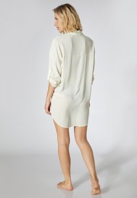 Crème-kleurige oversized shirtjurk met opgerolde mouwen, textuurstof en een ronde zoom, gezien vanaf de achterkant.