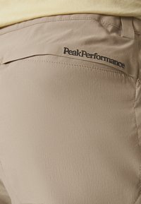 Pantalon beige en tissu lisse et léger. Dispose d'une poche arrière avec le logo "PeakPerformance" et des détails cousus.