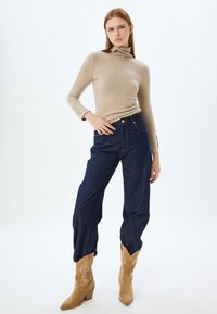 Maglione aderente beige con collo alto, jeans a vita alta blu scuro e stivali da cowboy in suede beige con dettagli cuciti e tacco spesso.