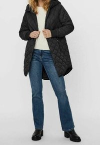 Manteau long matelassé noir avec capuche, présentant un col haut et des poches latérales, porté sur un pull crème et un jean bleu. Bottines noires.