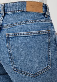 Calças de ganga azul com um remendo de couro bege rotulado "calças perfeitas" e detalhes em costura. O tecido parece texturizado com um design clássico.