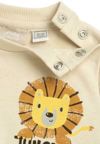 Body beige en coton avec un motif de lion. Comprend des boutons-pressions sur l'épaule pour un habillage facile. Texture douce, encolure ronde.