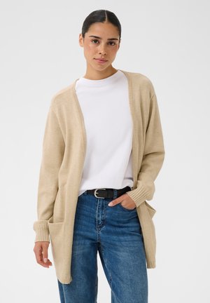 Femme aux cheveux foncés portant un cardigan beige, une chemise blanche et un jean bleu, debout avec une main dans la poche sur un fond clair uni.