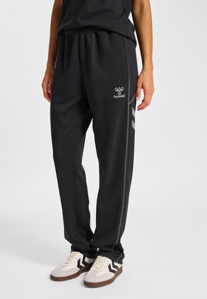 Personne portant un pantalon de survêtement Hummel noir avec un logo et des baskets blanches à rayures en zigzag noires, debout devant un fond uni.