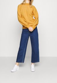 Person wearing mustard yellow sweater, wide-leg blue jeans, and white sneakers standing against plain white background.