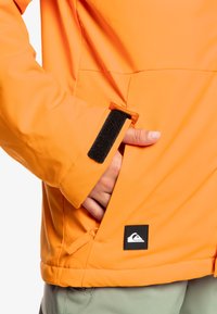Veste imperméable orange avec un poignet en velcro, une poche latérale et un patch logo noir sur l'ourlet inférieur. Texture lisse et coupe ajustée.