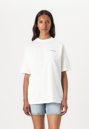 T-shirt bianco oversize in cotone morbido con collo giro. Presenta maniche corte e una piccola grafica discreta sul fronte. Indossato con pantaloni corti di jeans chiari.