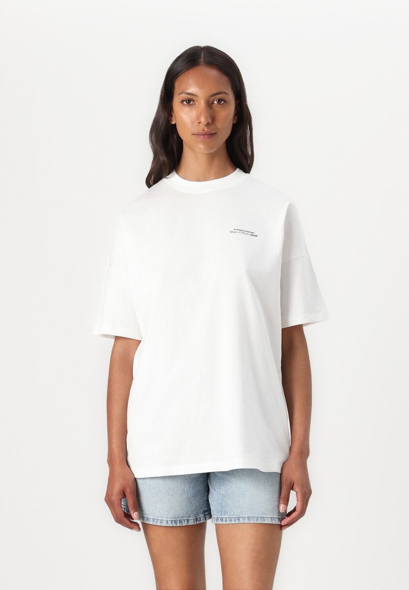 White oversized t-shirt in soft cotton with a crew neck. Features short sleeves and a small, subtle graphic on the front. Worn with light denim shorts.