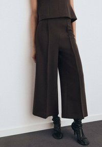 Pantalons larges marron en tissu lisse, avec une taille haute et des détails plissés, associés à des bottes à talons noires au design froncé.