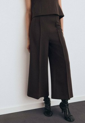 Pantalons larges marron en tissu lisse, avec une taille haute et des détails plissés, associés à des bottes à talons noires au design froncé.