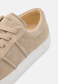 Sneaker in suede beige con suola in gomma bianca, dotato di lacci tono su tono e dettagli del marchio discreti sul lato. Texture liscia, design a profilo basso.