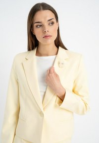 Jasnożółta marynarka o slim fit, z klapami typu notch i jednym guzikiem. Posiada teksturowany kwiatowy przypinek na lewym klapie.