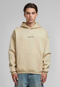 Beige katoenen hoodie met kangoeroezak en geribbelde manchetten. Heeft het logo "AnotherCotton" in het zwart, gecentreerd op de borst. Ontspannen pasvorm.