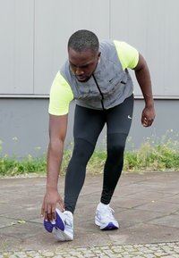 Athletischer Mann in eng anliegenden schwarzen Leggings und einem grauen Shirt mit neon-gelben Ärmeln, der sich auf dem Gehweg zu seinen weißen und lila Schuhen dehnt.