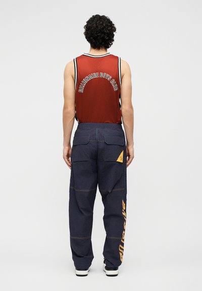 Άτομο, που φαίνεται από πίσω, φοράει κόκκινο αμάνικο μπλουζάκι "Billionaire Boys Club" και σκούρο μπλε παντελόνι cargo με κίτρινες λογότυπες στην τσέπη και στο πόδι.