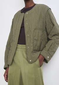 Kurtka w kolorze oliwkowym, pikowana, z zatrzaskami, okrągłym dekoltem i kieszeniami po bokach. Nosisz ją na ciemnym topie, w połączeniu z pasującymi jasnooliwkowymi spodniami.