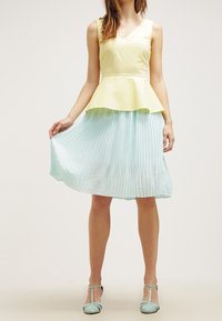Abito peplum con un corsetto giallo aderente e una gonna a pieghe verde menta. Presenta una scollatura a V e una texture di stoffa liscia. Un paio di scarpe color menta.