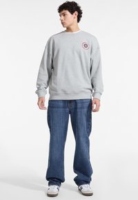 Felpa grigia con colletto rotondo e patch con logo, vestibilità comoda, polsini e orlo a coste; abbinata a jeans blu larghi e sneakers bianche.