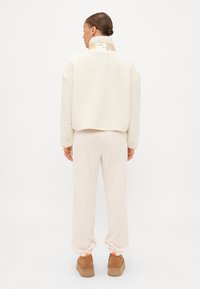 Jachetă din fleece crem cu un guler lucios, asortată cu pantaloni joggeri crem și încălțăminte maro. Prezintă un fit relaxat și mansete elastice.
