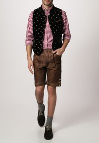 Mann in einer schwarzen, bestickten Samtweste, kariertem Shirt, braunen Lederhosen, grauen knielangen Socken und dunklen Schuhen.