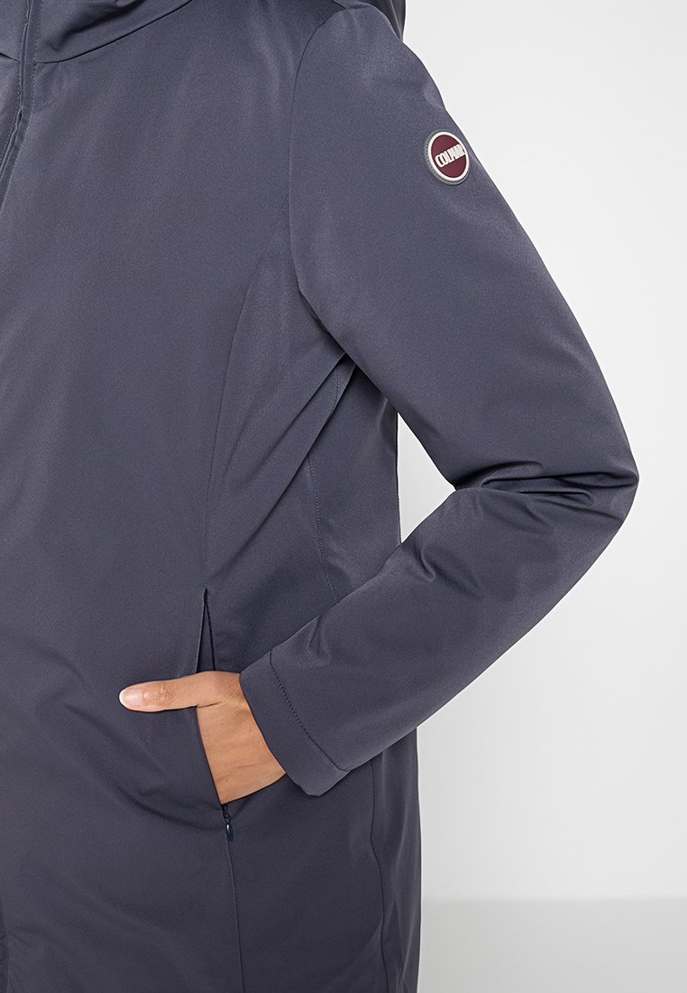 Marinblå jacka med en slät, vattenavvisande yta. Har sidofickor, dragkedjestängning och en cirkulär logotyp på vänster ärm.