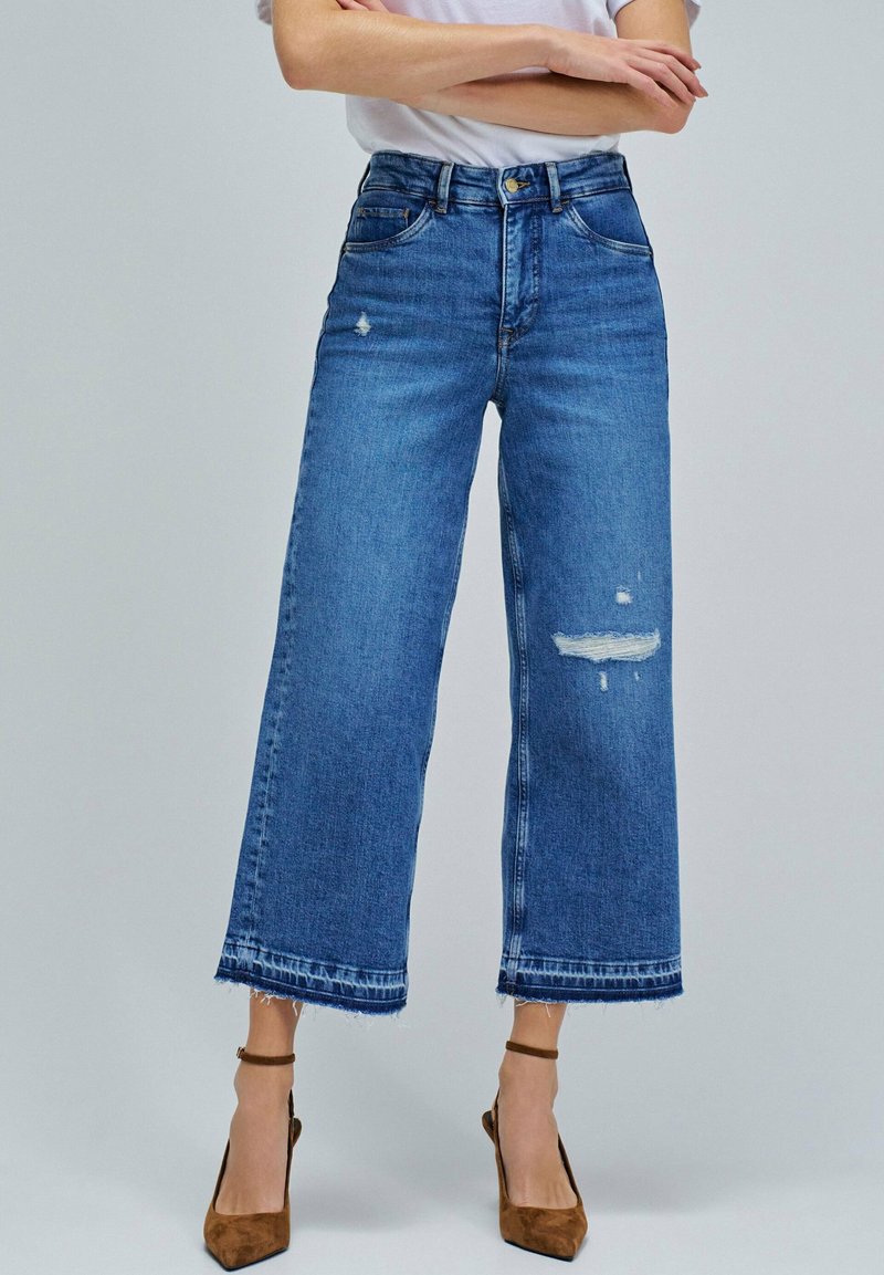 Vaqueros de mezclilla azul de pierna ancha con cintura alta, detalles desgastados y dobladillo deshilachado. Combinados con tacones marrones.