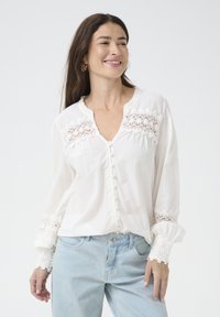 White button-down blouse with lace trim on shoulders and cuffs, featuring a v-neck design and slightly puffed sleeves, paired with light blue jeans.