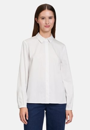 Hvid langærmet bluse med spids krave, knaplukning foran og elastiske manchetter, lavet af glat stof. Båret med mørkeblå jeans.